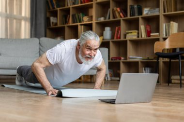 Spor kıyafetli yaşlı bir adam evde yoga minderinin üzerinde planking yapıyor dizüstü bilgisayarda fitness videosu izliyor, oturma odasının içinde, fotokopi odasında. Çevrimiçi fitness sınıfı, yaşlılar için spor yaşam tarzı