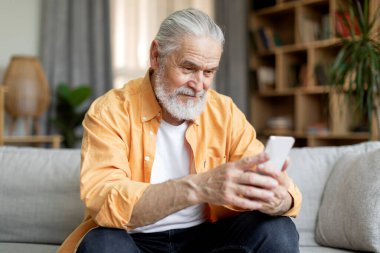 Happy handsome senior man sitting on couch at home, using modern smartphone, checking social media while chilling alone at weekend, copy space. Leisure and gadgets for seniors