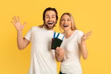 Glad happy excited millennial caucasian man and lady in white t-shirts with open mouths showing passports with air tickets, isolated on yellow background. Travel, vacation, trip and facial expressions