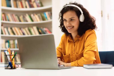 Happy mixed race woman in headphones studying online with laptop in library interior, watching webinar, enjoying distance learning and smiling, free copy space