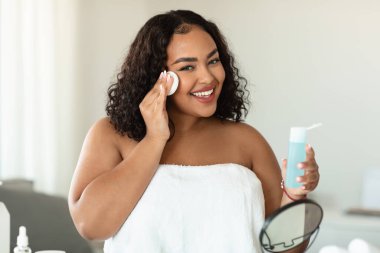 Beautiful black chubby lady cleaning her face, using cotton pads and cleansing product, looking at camera and smiling. Young lady using face toner and cotton pad, home interior