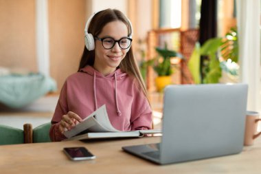 E-learning, web-based education concept. Happy teen girl studying online from home, reading book and looking at laptop pc, sitting at desk, free space