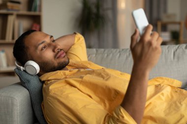 Attractive young black guy in headphones listening to music online on mobile phone, lying on sofa at home. Handsome millennial African American man in headset watching video on cellphone