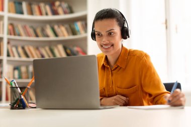 Happy female student learning online with laptop, having remote class and taking notes, lady wearing headphones and smiling at screen, sitting in library. Modern education and e-learning concept