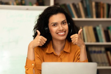 Online teacher. Happy mixed race female tutor in headphones showing thumbs up, looking and smiling at camera while sitting in library and having online classes with students