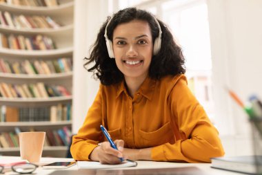 Happy woman in headset having online lesson on laptop, writing in notebook and smiling at camera, sitting in modern library. Female student taking notes during webinar, studying remotely