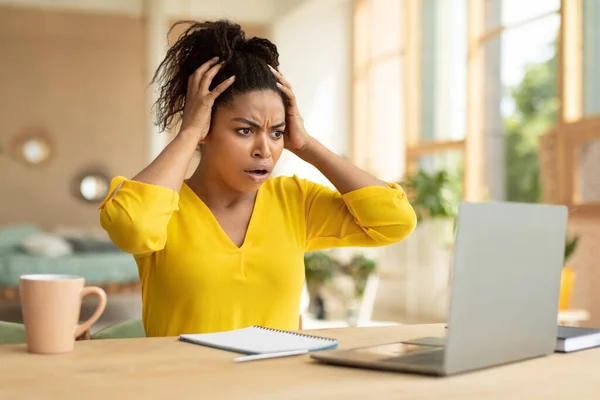 Oh no, shocking business news concept. Emotional african american businesswoman looking at laptop in shock and touching head, having problem, sitting at desk at home