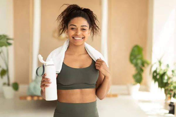 Nutrition for sport and exercise. Happy african american woman with white towel on neck holding shaker with protein, smiling at camera after domestic workout