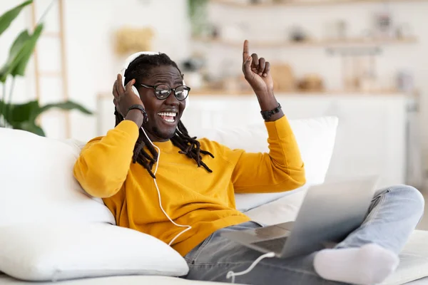 Cheerful Black Guy Wearing Headphones Relaxing On Couch And Listening Music On Laptop, Positive Young African American Man Having Fun At Home, Dancing While Enjoying Favorite Playlist, Copy Space