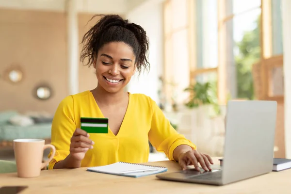 Making financial transaction. Excited black lady holding credit card and using personal computer, sitting at desk at home. Online shopping and payment concept