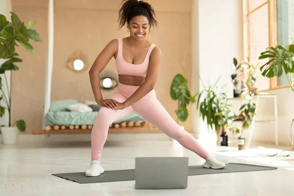 Sporty african american lady exercising at home, stretching her legs, watching online tutorial on laptop or having online fitness class, bedroom interior, copy space