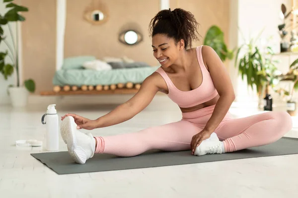 Happy fit black lady in sportswear exercising at home, stretching on fitness mat, touching her feet with hand, cozy bedroom interior, copy space