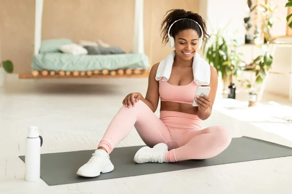 Happy black fitness lady using smartphone and wearing headphones, having break during domestic workout, sitting on yoga mat at home. Sport and technology concept
