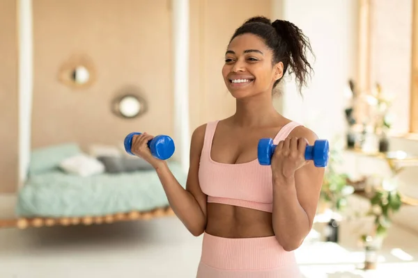 Portrait of happy african american lady doing dumbbell workout at home, working on arms strength and smiling, copy space. Fit woman lifting blue fitness dumbells up over bedroom interior