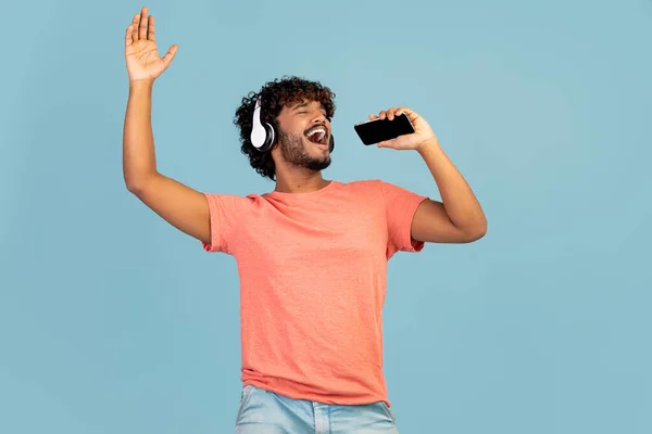 Joyful millennial indian guy singing karaoke and gesturing, using nice mobile application on newest smartphone and wireless headset, blue studio background, mockup, copy space
