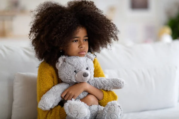 Upset preteen african american girl with curly bushy hair sitting on couch alone, embracing teddy bear, looking at copy space. Kid feeling lonely at home, missing parents or friends