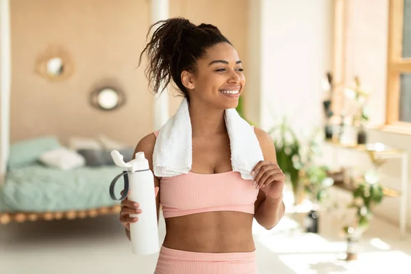 Smiling african american lady in sportswear having break while exercising at home, holding bottle with water and touching towel on her shoulders, copy space