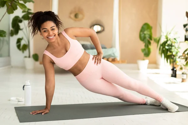 Happy young african american lady standing in side plank on yoga mat and smiling at camera, working out at home. Fit black woman in sports outfit exercising core muscles, doing strength workout