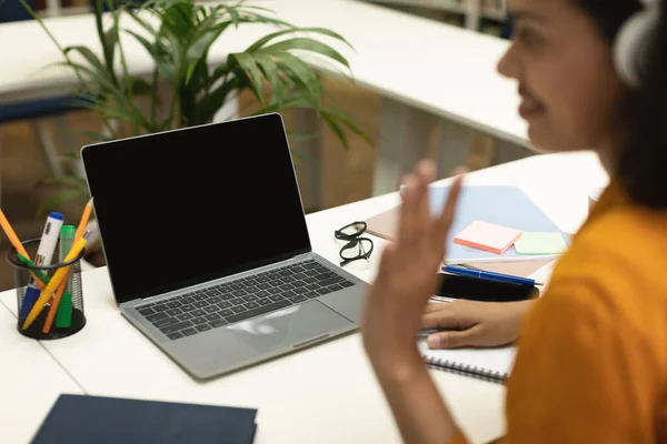 Video call. Black teacher waving hand at laptop blank screen, communicating online sitting at table in library. Modern distant communication, mockup