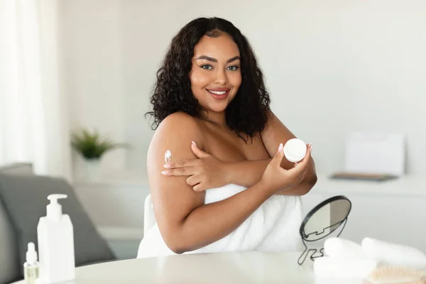 Skin care. Pretty black bodypositive woman applying moisturising body lotion after bath, standing with cosmetic jar in hand in bathroom interior