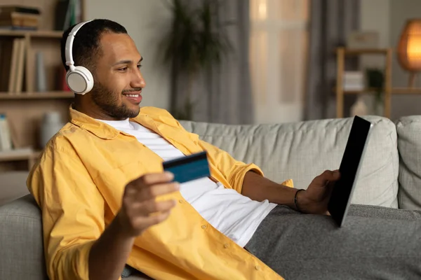 Cheerful Black Man Laptop Credit Card Lying Couch Wearing Headphones — 스톡 사진