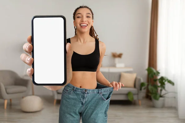 Weight Loss App. Happy Slim African American Woman Wearing Large Jeans Showing Blank Smartphone With White Screen, Smiling Black Female Recommending Dieting Application, Mockup, Collage