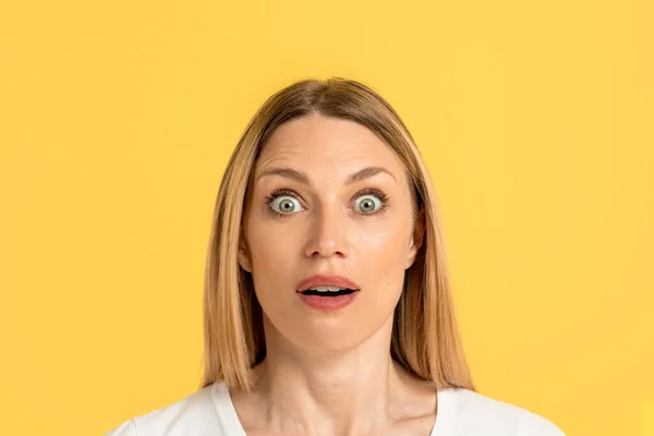 Cheerful surprised millennial caucasian blonde lady with open mouth in white t-shirt looks at camera, isolated on yellow background, close up. Shocked ad and amazed offer, human facial expression