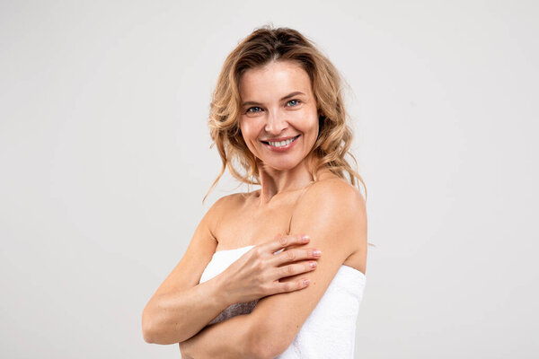 Natural Beauty. Portrait of cheerful middle aged female with beautiful skin standing wrapped in bath towel over light studio background, attractive woman enjoying self-care routine, free space