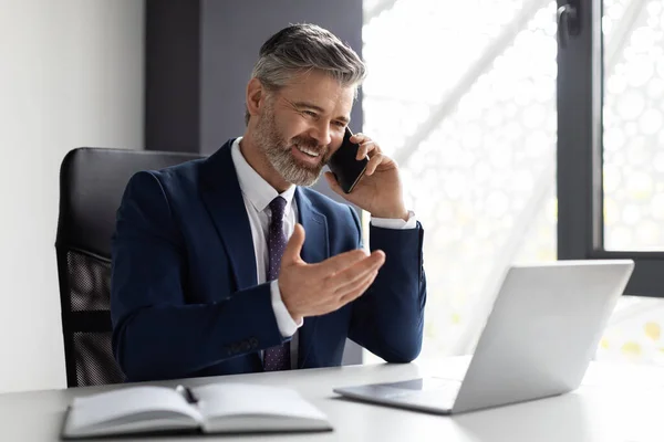 Corporate Communication. Handsome Middle Eaged Businessman Talking On Cellphone In Office, Smiling Male Entrepreneur In Suit Having Mobile Conversation While Sitting At Desk With Laptop, Copy Space