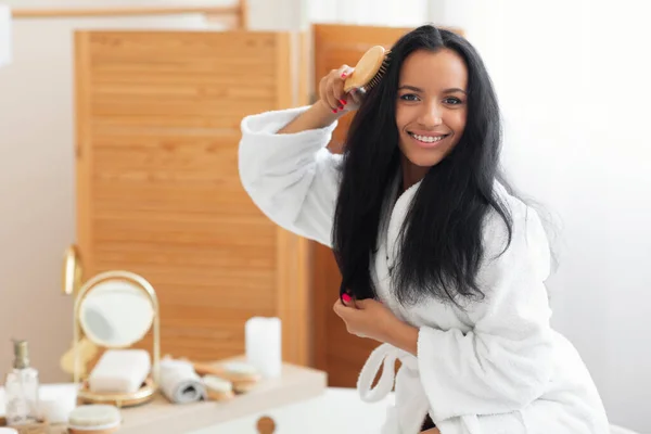 Happy Mixed Woman Brushing Hair Posing With Hairbrush Smiling To Camera In Modern Bathroom Indoor. Pretty Lady Detangling Long Brunette Hair. Haircare Concept