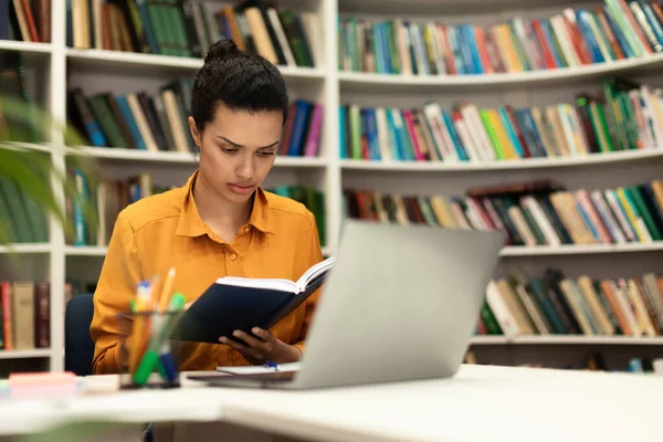 Concentrated Mixed Race Woman Reading Book Sitting Library Laptop Study — Stockfoto