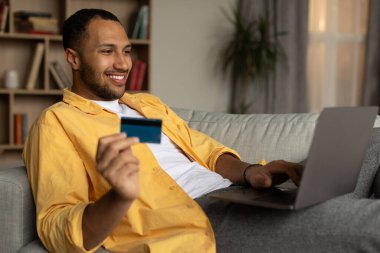 Remote shopping concept. Cheerful African American man in headphones buying things on web, using laptop and credit card in living room. Positive young black guy ordering goods online from home