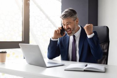 Joyful Middle Aged Businessman With Cellphone And Laptop Celebrating Success In Office, Happy Male Entrepreneur In Suit Looking At Computer Screen, Shaking Fists And Exclaiming With Excitement