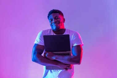 Cheerful young black man hugging laptop with empty screen in neon light, mockup for website. Tech nerd embracing portable pc, offering copy space for your ad. Technology lover concept