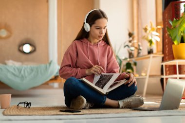 Focused caucasian teen girl sitting in wireless headphones and holding book, looking at laptop screen in bedroom interior at home, copy space