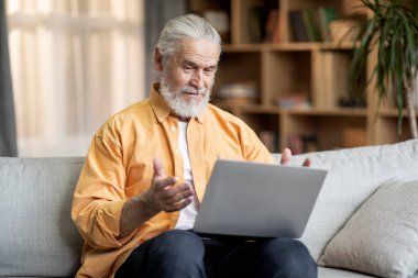 Cheerful bearded senior man in casual outfit sitting on couch at home, having online party with family, using computer, looking at laptop screen, smiling and gesturing, copy space