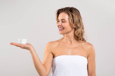 Beauty Care For Aged Skin. Attractive Middle Aged Woman Holding Jar With Face Cream On Her Palm, Smiling Beautiful Female Wrapped In Bath Towel Advertising New Cosmetics, Standing On Grey Background