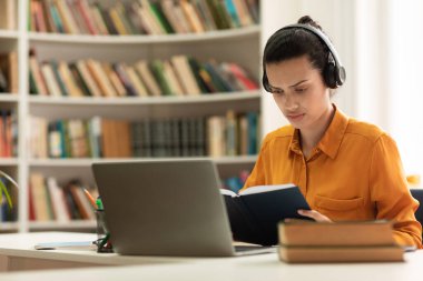 Focused female student reading book and using laptop computer, studying online, sitting in library, copy space. Remote education, distance learning and exam preparation concept