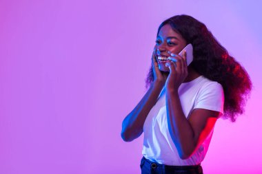 Charming young black woman speaking on cellphone, having phone conversation in neon light, empty space. Millennial African American female communicating on smartphone