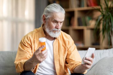 Concentrated grey-haired bearded senior man sitting on couch at home, holding yellow jar with medicine and modern smartphone, checking information about drugs on Internet, copy space