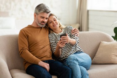 Happy Middle Aged Couple Using Mobile Phone Texting And Browsing Internet Sitting On Sofa At Home. Husband And Wife Posing With Cellphone. New Application, Communication Concept