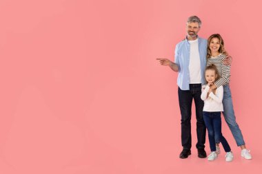 Happy Parents And Kid Daughter Pointing Finger Aside Recommending Offer, Showing Copy Space Standing On Pink Studio Background. Family Advertising Your Text. Full Length