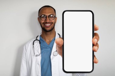 Cheerful attractive young arabic man practitioner showing newest smartphone with white empty screen and smiling, recommending brand new application for healthcare, white background, mockup