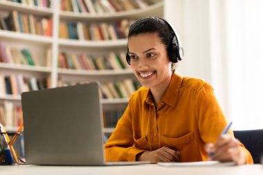 Happy mixed race lady having online class, using headset, looking at laptop screen and taking notes, sitting in library. E-education, webinar, online course concept
