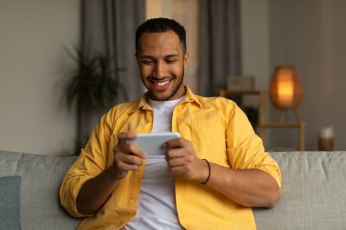 Happy young black man using smartphone, chatting online, sitting on couch at home. Portrait of millennial African American guy communicating on webcam via mobile device, indoors