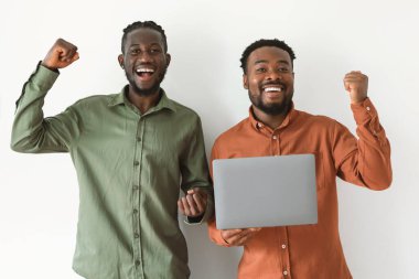 Two Joyful African American Men Holding Laptop Shaking Fists Celebrating Online Business Success Smiling To Camera Standing Posing Over White Studio Background. Victory Celebration