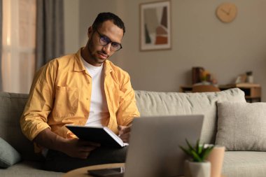 Focused young black man using laptop pc, working or studying online, taking notes during business meeting or webinar, having remote lesson, sitting on couch at home. Distance work, education concept