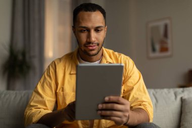 Focused young black man with tablet sitting on sofa, working or studying online, having remote business meeting at home. Serious African American guy communicating on web via touch pad