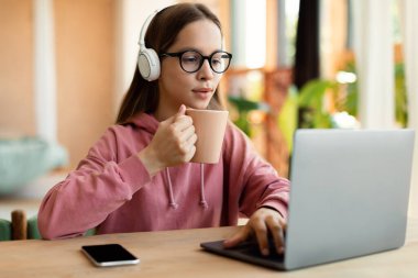 Teenager girl having online lecture on laptop at home, wearing wireless headset and holding cup of tea. Focused youth having webinar or conference on pc, typing on computer keyboard