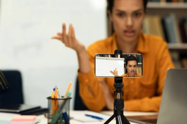 Young female teacher providing online lesson looking at cellphone camera or recording video on smartphone on tripod, explaining new material to students